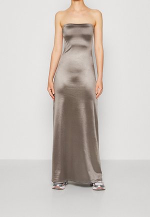 Femme portant une robe grise en satin, sans bretelles et longue jusqu'au sol, associée à des baskets argentées et blanches, debout contre un mur uni.