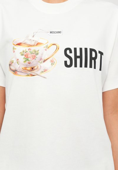 Tricou alb din bumbac cu un imprimeu al unei cești de ceai florale, având un plic de ceai atașat și cuvântul „TRICOU” scris cu litere negre îngroșate.
