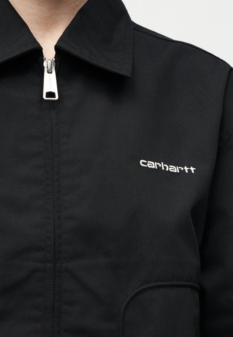 Primo piano di una giacca nera con zip e colletto, con logo "carhartt" ricamato in bianco sul petto.