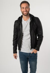 Giacca di pelle nera con zip frontale e cappuccio attaccato, caratterizzata da una finitura testurizzata. Abbinata a una maglietta chiara e jeans blu.