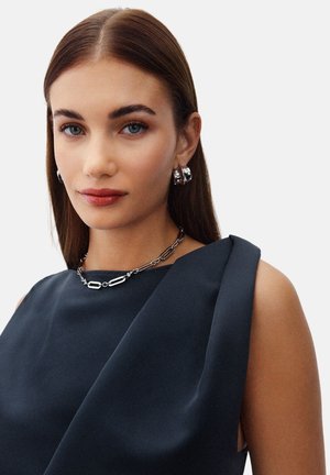 Abito nero senza maniche con una texture liscia, caratterizzato da un design asimmetrico sulle spalle. Una collana di catena argentata e orecchini a cerchio completano il look.
