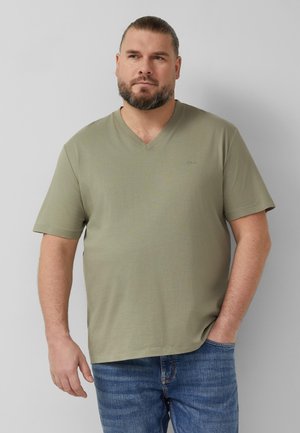 Bearded man with tied-back hair wearing a light olive green V-neck T-shirt and blue jeans, standing against a plain gray background.