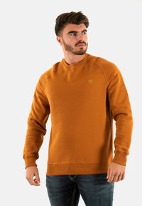 Sudadera de cuello redondo en color ámbar con puños y dobladillo de canalé. Presenta un logo en el pecho. Fabricada con un tejido suave y de textura lisa.
