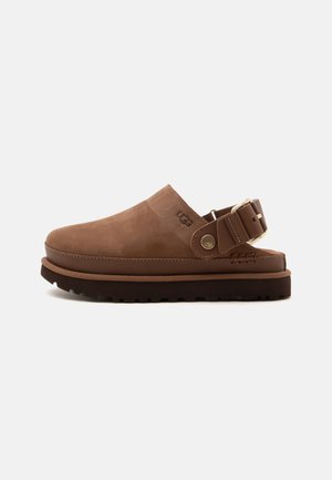 Sabot marron à enfiler avec une bride boucle ajustable, semelle noire épaisse et logo UGG sur le côté, présenté de profil sur fond blanc.