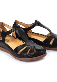 Les sandales en cuir noir présentent des découpes, un intérieur en cuir beige et des semelles en caoutchouc marron avec des coutures détaillées le long des bords.