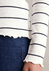 Haut à manches longues blanc à rayures horizontales bleu marine, avec un ourlet festonné et une petite étiquette logo sur le côté. Porté avec un jean bleu foncé.