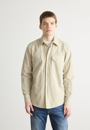 Beige long-sleeve shirt in cotton, featuring a button-down collar, chest pocket, and light texture, paired with blue denim jeans.
