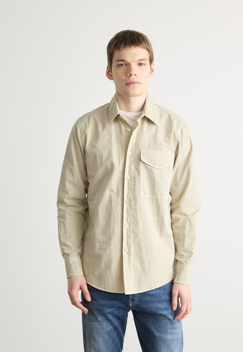 Camicia beige a maniche lunghe in cotone, dotata di colletto con bottoni, tasca sul petto e texture leggera, abbinata a jeans in denim blu.