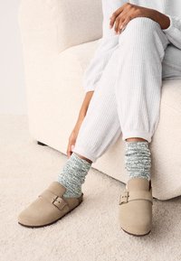 Sabots en daim beige à bout arrondi avec une boucle ajustable, associés à des chaussettes texturées côtelées vertes et blanches, portés avec une tenue décontractée gris clair.