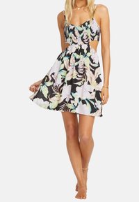 Frau in einem ärmellosen, floralen Minikleid mit Ausschnitten an der Taille, die barfuß steht und den Rock leicht anhebt.