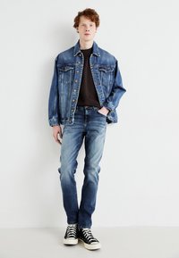 Jachetă din denim cu spălare deschisă, două buzunare la piept, închidere cu buton; combinată cu un tricou negru și blugi albaștri ușor decolorați, teniși negri.
