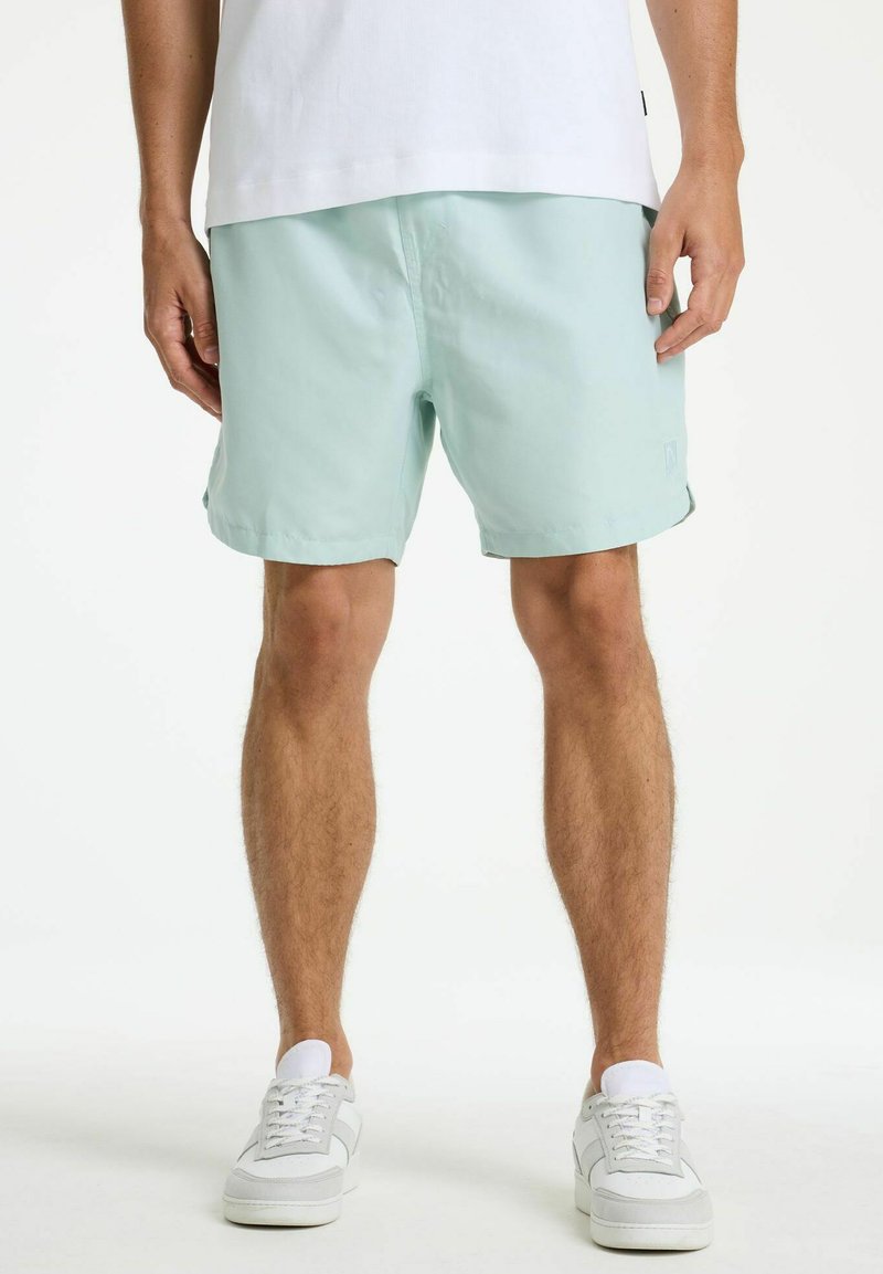 Uomo con pantaloncini verde chiaro, scarpe da ginnastica bianche e camicia bianca, in piedi con le braccia rilassate lungo i fianchi.