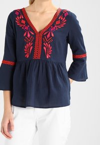 Blusa blu navy con scollatura a V, motivi floreali ricamati in rosso e rosa, maniche svasate e vita arricciata.