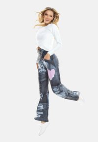 Sieviete lec plašās biksēs ar sirds rakstu un rozā tonējošu garroku T-kreklu, smaidot, uz vienkrāsaina baltā fona.