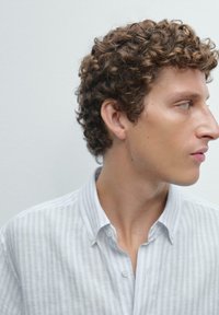Chemise à col rayée bleu clair et blanche, fabriquée dans un tissu doux. Dotée de boutons et d'une coupe décontractée. Le modèle a des cheveux bouclés.