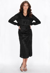 Schwarzes Satinkleid mit langen Ärmeln, durchgehender Knopfleiste, Raffungsdetails und seitlichem Schlitz. Kombiniert mit silbernen Riemchen-High Heels.