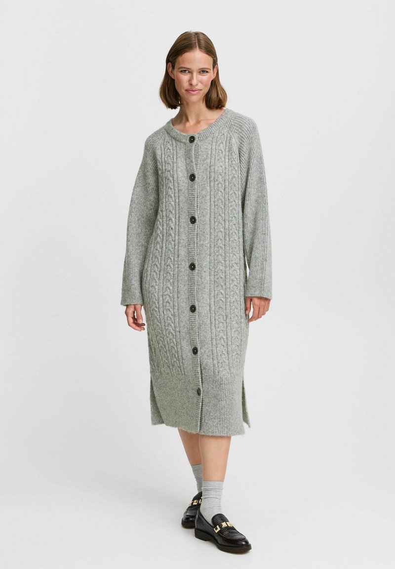 Vestido de cárdigan gris de punto que presenta patrones de trenzado, cierres de botones, mangas largas y aberturas en los laterales, elaborado con una suave mezcla de lana texturizada.