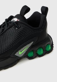 Sapatilhas pretas com parte superior em malha texturizada, detalhes em verde de amortecimento de ar, acabamentos refletivos e um padrão geométrico distintivo na lateral.