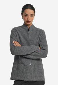 Szary, fakturowany żakiet z kołnierzykiem mandaryńskim, zapięciem na guziki z przodu i dwoma kieszeniami naszytymi. Wykonany z tkaniny plecionej o subtelnym wzorze.