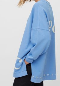 Ljusblå oversized sweatshirt gjord av mjuk bomull, med en vit logotryck och nedhasade axlar. Sömmar vid ärmarna.