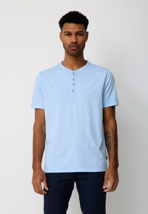 Mann mit kurzen lockigen Haaren, der ein hellblaues Henley-Shirt mit kurzen Ärmeln und eine dunkle Hose trägt, stehend vor einem schlichten weißen Hintergrund.