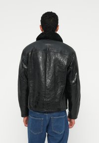 Jaqueta preta de couro sintético com um acabamento texturizado e um colarinho de pele de carneiro preta. Combinada com jeans de denim azul. Detalhes de costura visíveis.