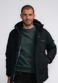 Schwarze isolierte Jacke mit Reißverschluss und Druckknopfverschluss, ausgestattet mit einer Kapuze und mehreren Taschen. Tragen über einem grünen Pullover.
