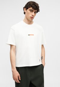 Νέος άνδρας με σγουρά μαλλιά φοράει λευκό t-shirt Nike με το λογότυπο "FUBBALL" και μαύρο παντελόνι, στέκεται μπροστά σε απλό φωτεινό φόντο.
