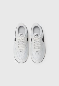 Par laka bijelih Nike Air Force 1 tenisica s crnim Swoosh logotipovima, bijelim vezicama i perforiranim prednjim dijelovima na običnoj bijeloj pozadini.