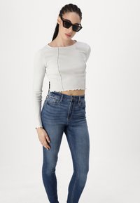 Λευκή ριγωτή μπλούζα μακρύς μανίκι με κοντό στρίφωμα, συνδυασμένη με τζιν ψηλής μέσης σε μπλε χρώμα. Στρογγυλή λαιμόκοψη και διακριτές φθορές στις άκρες.