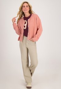 Roze pluizige cardigan met knopen aan de voorkant, gedragen over een bordeauxrood shirt, gecombineerd met beige broek en crèmekleurige sneakers. Natuurlijke omgeving.