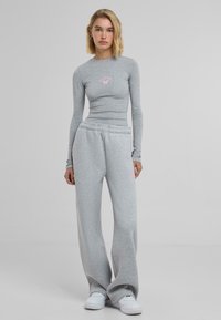 Grå langermet topp med en tettsittende design og rosa grafikk nær brystet, kombinert med løst sittende grå joggebukser og hvite joggesko.