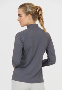 Dunkelgraues, eng anliegendes Sportoberteil mit langen Ärmeln, hergestellt aus einem glatten, dehnbaren Material. Verfügt über einen hohen Kragen und Raglanärmel für optimale Bewegungsfreiheit.