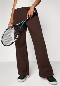 Brown, wide-leg athletic pants made of soft fabric, paired with a black and blue tennis racket and black athletic shoes with textured detailing.