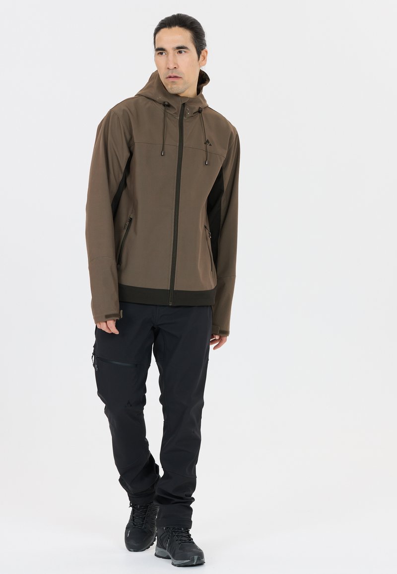 Braune wasserdichte Jacke mit Kapuze, mit schwarzen Seitenpaneelen, Reißverschlusstaschen und taillierter Passform, kombiniert mit schwarzen Hosen und Stiefeln.