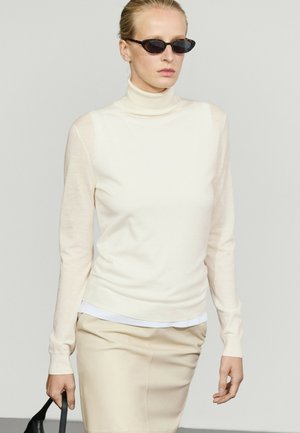 Femme portant un pull à col roulé crème et une jupe beige, tenant un sac noir, avec des lunettes de soleil ovales foncées et les cheveux tirés en arrière.