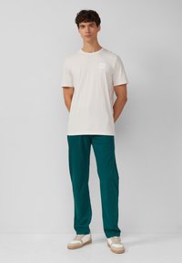 Lichtbeige katoenen t-shirt met een ronde hals, voorzien van een klein logo aan de linkerkant, gecombineerd met teal kleurige broeken en beige sneakers.