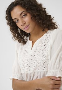 Blusa bianca con dettagli in pizzo, caratterizzata da un motivo a zigzag, maniche corte a palloncino e scollatura arrotondata. Tessuto morbido e leggero.