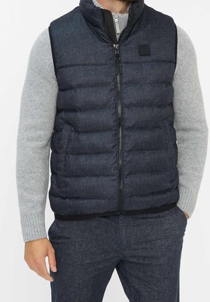 Mężczyzna ubrany w ciemnoniebieską pikowaną kamizelkę bez rękawów na jasnoszarym swetrze z długim rękawem, w ciemne, kraciaste spodnie, z rozluźnionymi rękami.
