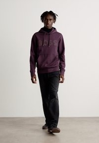 Lila Kapuzenpullover mit "BOSS"-Logo, Fronttasche und Zugkordelkapuze, kombiniert mit schwarzen Hosen und dunklen Schuhen vor einem minimalistischen Hintergrund.