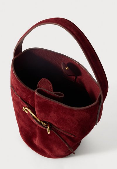 Ümar burgundi sametist ämbritasku, millel on õhuke nahkrihm, kuldsete detailidega, nööbiklõps ja pehme, tekstureeritud sisepolster.