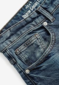 Jeans din denim albastru închis, cu un design clasic cu cinci buzunare, cu cusături vizibile și un detaliu metalic la talie.