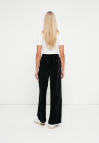 Vrouw met lang blond haar, gekleed in een wit shirt met korte mouwen, zwarte broek met wijde pijpen en witte sneakers, staande met haar gezicht naar een eenvoudige witte muur.