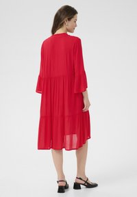 Robe rouge à étages en tissu léger avec un col à volants et de longues manches évasées. Portée avec des chaussures à talons noires.