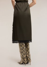 Olivgrön satinklänning med svart spetskant, kombinerad med figursydda byxor i ormskinnsmönster i beige och bruna nyanser, med spetsiga klackar.