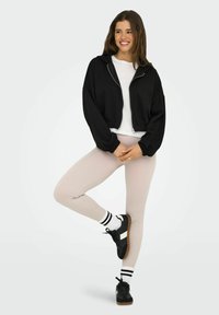 Hoodie noir court avec zip, chemise blanche, leggings beiges côtelés, baskets noires avec bande blanche et chaussettes rayées en noir et blanc.