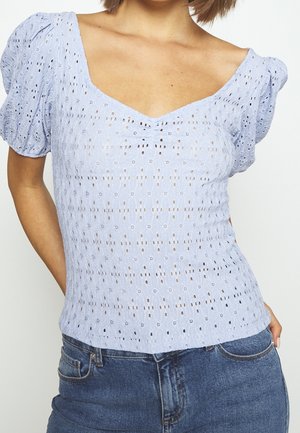 Lyseblå top med puffærmer, lavet af tekstureret stof med hulmønstre. V-udskæring, tætsiddende silhuet, parret med denim.