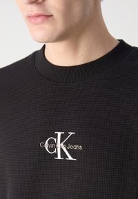 Fekete, texturált kapucnis pulóver ribbelt nyakkal, fehér és világos bézs hímzett "Calvin Klein Jeans" logóval a mellrészen.