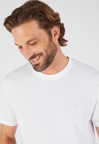 Camiseta blanca de manga corta con textura acanalada y cuello redondo, que presenta un discreto logo bordado en el pecho.