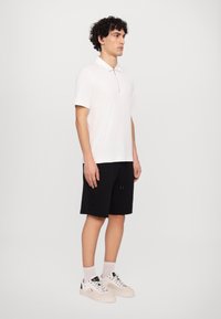 Tricou polo alb cu mânecă scurtă, guler și fermoar, asortat cu pantaloni scurți negri până la genunchi și adidași albi cu accente negre.